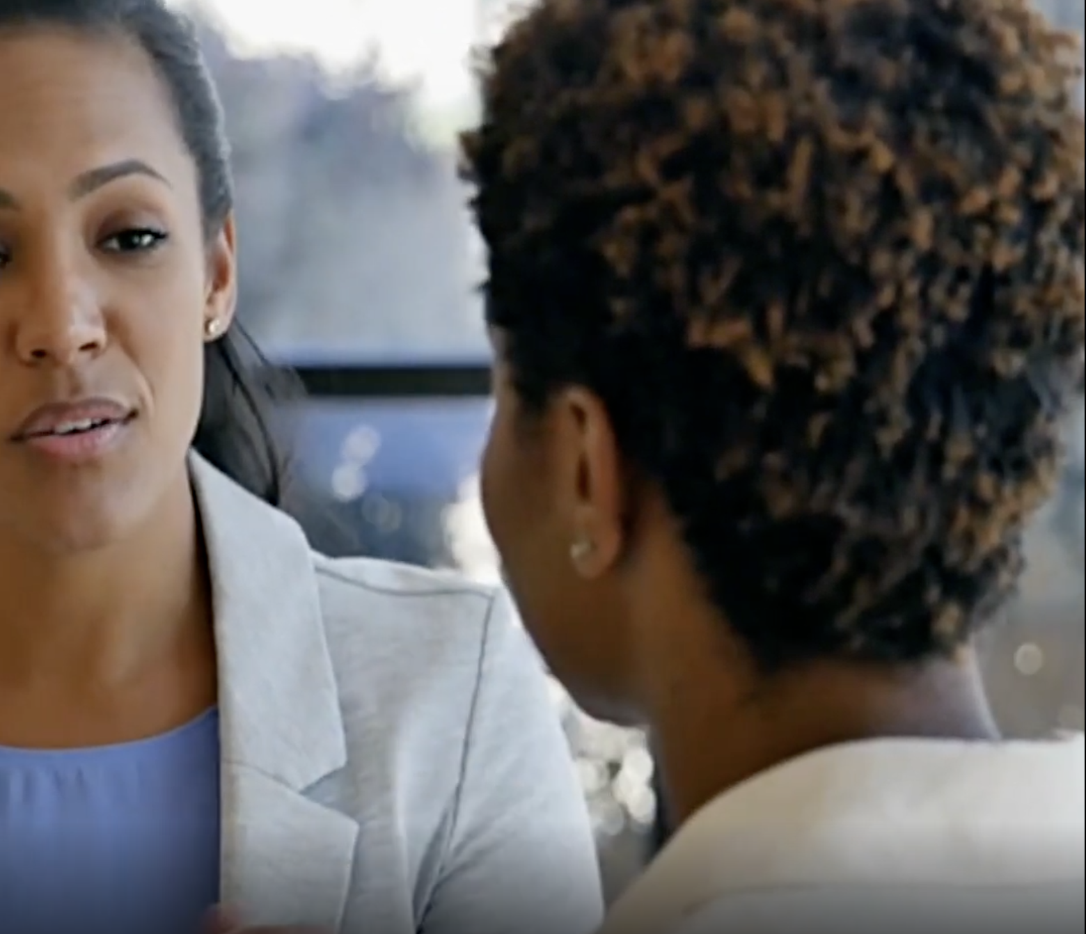 Two women are seated and talking indoors, with one woman facing the camera and the other shown from behind.