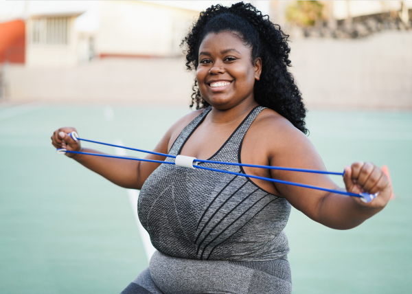 A woman in athletic wear smiles while exercising outdoors, holding and stretching a blue resistance band with both hands.