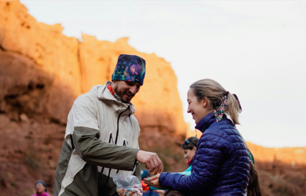 Two people exchange snacks outdoors, dressed in jackets and headwear, with a rocky, sunlit cliff in the background.
