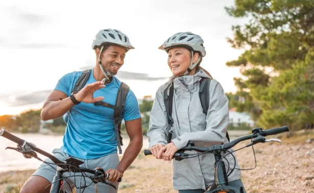 Two people wearing helmets and sportswear are standing outdoors with their bicycles, smiling and talking near a body of water and trees.