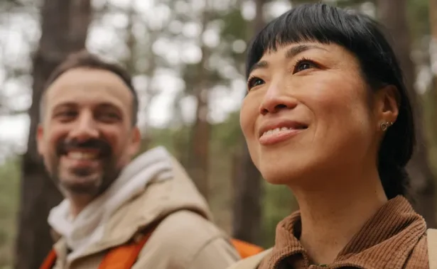 Two people outdoors in a wooded area, both wearing jackets. The person in the foreground is smiling and looking up; the person in the background is slightly blurred and also smiling.