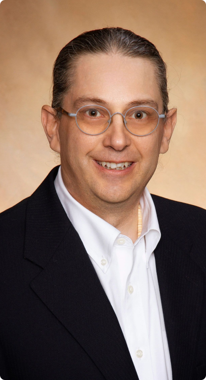 A person with glasses and long hair pulled back, wearing a dark blazer and white collared shirt, poses in front of a plain beige background.