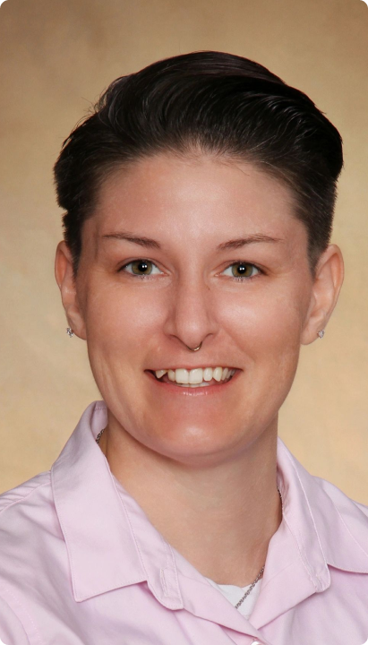 A person with short dark hair, light skin, and green eyes is smiling. They are wearing a light pink collared shirt and small earrings, in front of a beige background.
