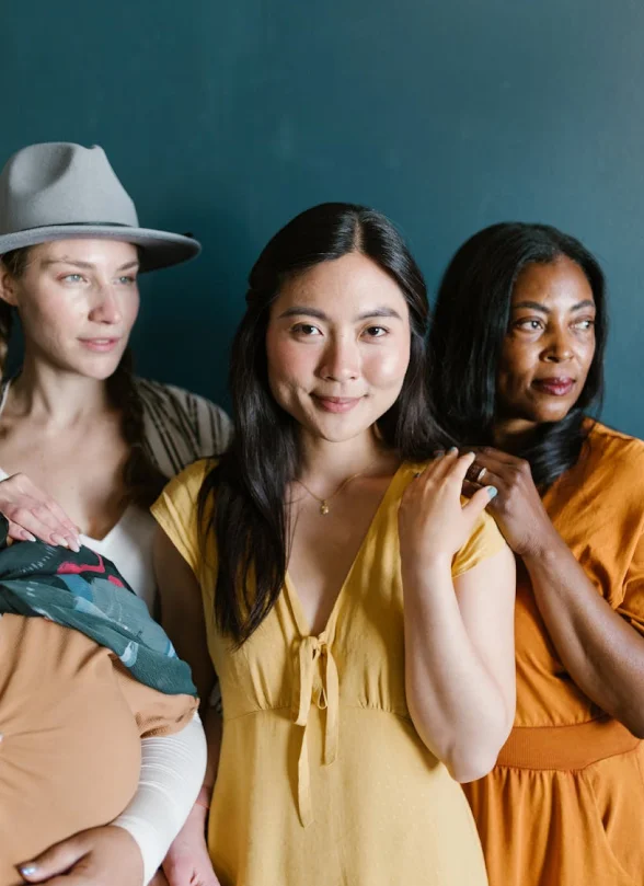 Three women stand together in front of a dark teal wall. The woman in the center wears yellow, the one on the left wears a hat, and the one on the right wears an orange dress.