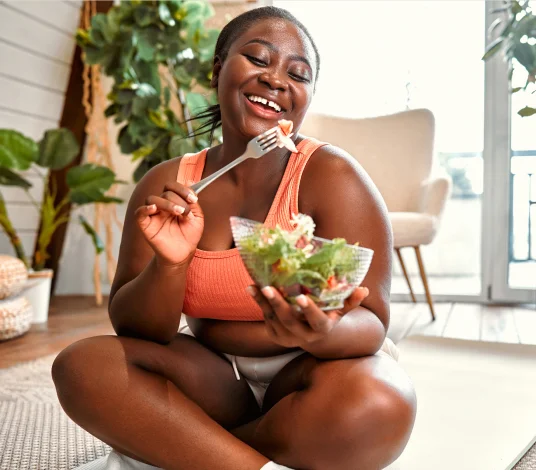 A woman in athletic wear sits on the floor indoors, smiling while eating a salad from a bowl with a fork. Plants and a chair are visible in the background.