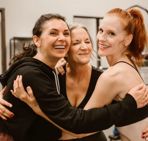 Three women are standing closely together indoors, smiling and embracing each other in a friendly, happy manner.