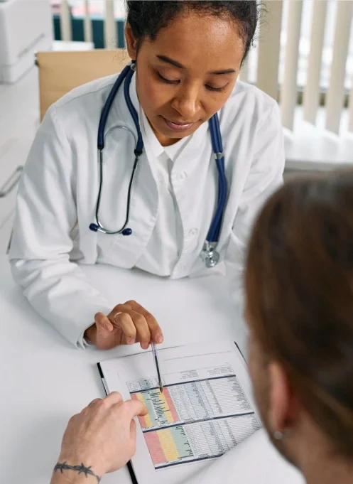 A doctor in a white coat uses a pen to explain a medical chart with colored sections to a patient across the desk.