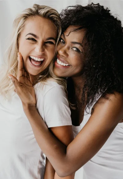 Two women smiling and embracing closely, both wearing white shirts against a light background.
