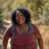 A woman with curly hair, wearing a maroon shirt and backpack, smiles while hiking on a sunlit forest trail.